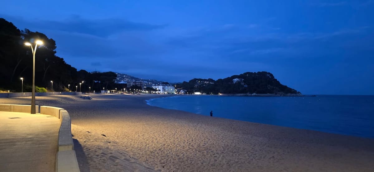 Lloret de Mar beach at dusk, empty sand and a lit promenade lamp with the town on the headland